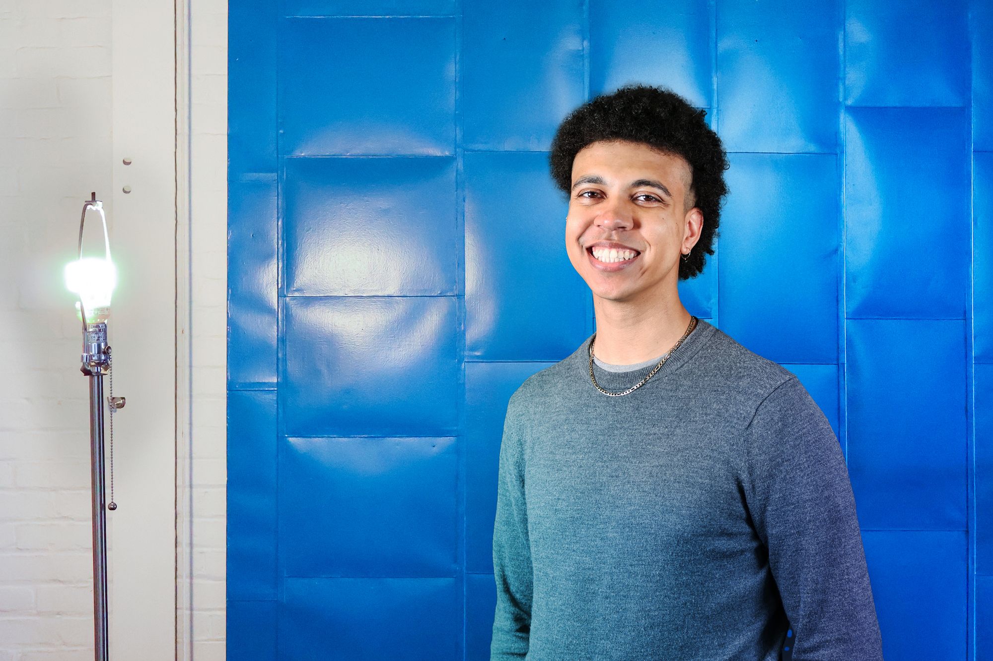 Young man smiling in front of a bright blue, padded wall. He is wearing a gray long-sleeve shirt and a silver chain necklace. A lit floor lamp stands to the left, casting a soft glow in the otherwise minimal, indoor setting.